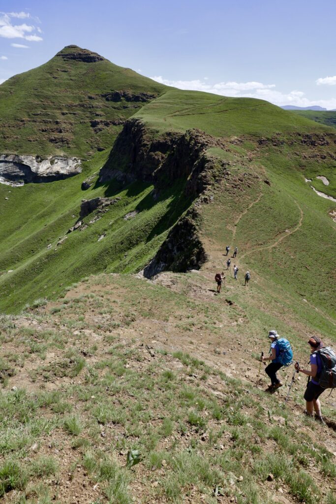 Des randonneurs munis de sacs à dos et de bâtons de trekking marchent sur un sentier de crête herbeux et accidenté dans un paysage montagneux sous un ciel bleu clair, avec une colline abrupte et des falaises en arrière-plan. - Karavaniers