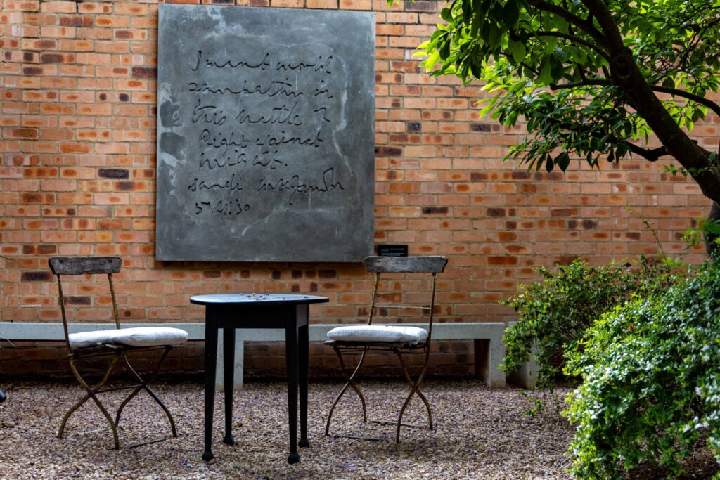 Deux chaises en métal avec des coussins et une table ronde noire sont assises sur du gravier dans un jardin extérieur. Un arbre et de la verdure entourent l'endroit. Une grande plaque grise avec une écriture cursive est accrochée à un mur de briques à l'arrière-plan. - Karavaniers