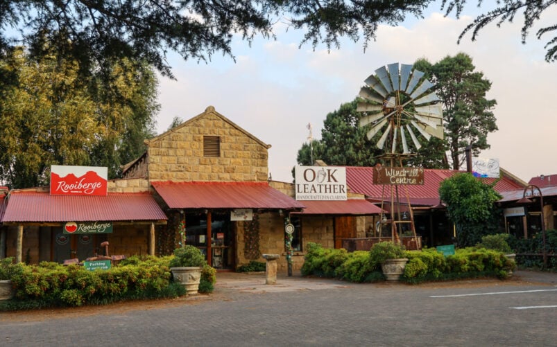 Une rangée de boutiques rustiques avec des façades en pierre et en bois, des toits rouges et des enseignes visibles telles que "Roseberge" et "O.K. Leather". Un moulin à vent et de la verdure sont présents devant les bâtiments. - Karavaniers