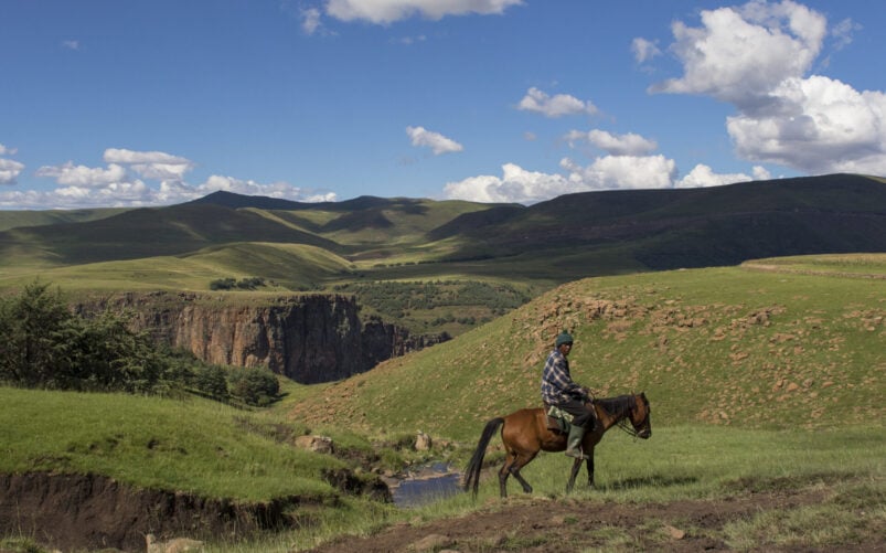 Une personne traverse à cheval un paysage herbeux composé de collines ondulantes, de falaises rocheuses et d'un ruisseau sous un ciel partiellement nuageux. - Karavaniers