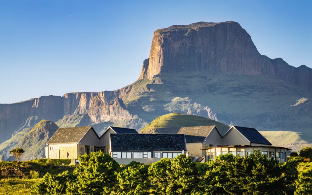 Un lodge moderne avec de grandes fenêtres et des toits en pente se dresse devant une montagne spectaculaire aux falaises abruptes, entourée de collines et d'arbres verts sous un ciel bleu clair. - Karavaniers