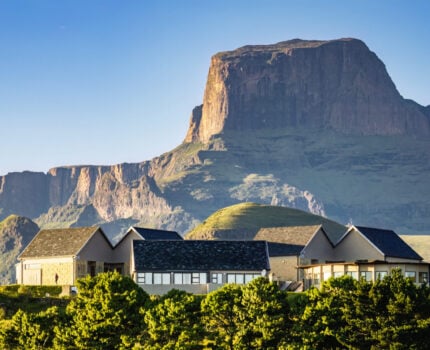 Un lodge moderne avec de grandes fenêtres et des toits en pente se dresse devant une montagne spectaculaire aux falaises abruptes, entourée de collines et d'arbres verts sous un ciel bleu clair. - Karavaniers