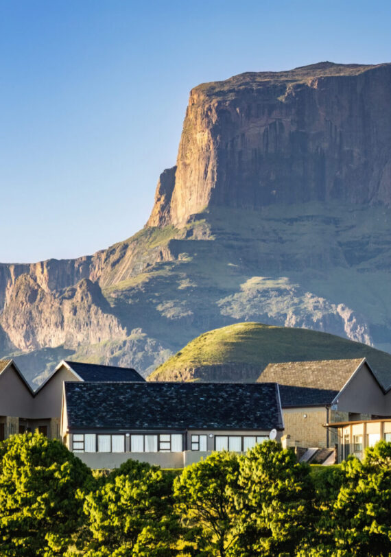 Un lodge moderne avec de grandes fenêtres et des toits en pente se dresse devant une montagne spectaculaire aux falaises abruptes, entourée de collines et d'arbres verts sous un ciel bleu clair. - Karavaniers