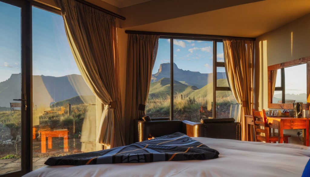 Une chambre à coucher avec de grandes fenêtres et portes vitrées offre une vue sur les montagnes vertes et le ciel bleu. La lumière du soleil entre à flots et éclaire un lit fait, une chaise, un bureau et une couverture pliée au pied du lit. - Karavaniers