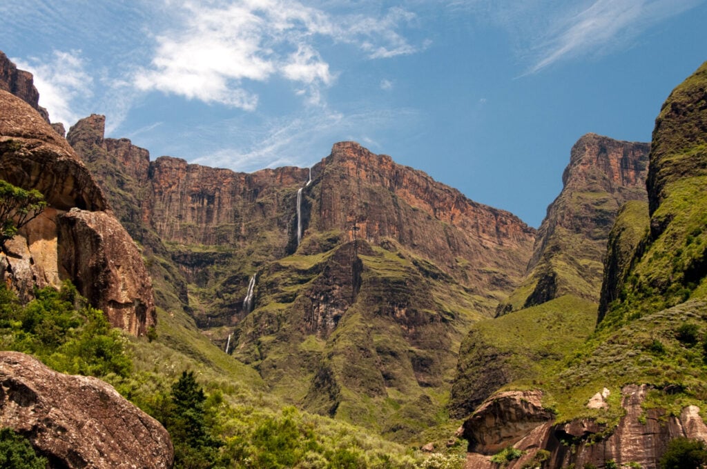 Une cascade haute et étroite se déverse sur le flanc d'une falaise accidentée de couleur brun-rouge dans un paysage montagneux verdoyant, sous un ciel bleu partiellement nuageux - un spectacle inoubliable lors d'un trek en Afrique du Sud. - Karavaniers