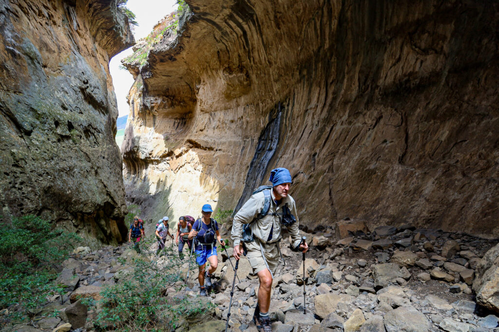Un groupe de randonneurs munis de bâtons de trekking remonte un sentier de canyon étroit et rocailleux entre de hautes parois rocheuses abruptes, lors d'un trek en Afrique du Sud. La végétation est rare et la lumière du soleil éclaire une partie du canyon. - Karavaniers