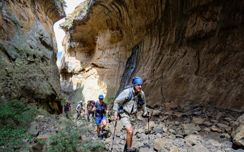 Un groupe de randonneurs munis de bâtons de trekking remonte un sentier de canyon étroit et rocailleux entre de hautes parois rocheuses abruptes, lors d'un trek en Afrique du Sud. La végétation est rare et la lumière du soleil éclaire une partie du canyon. - Karavaniers