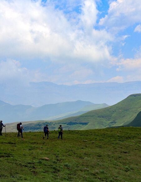 Un groupe de sept randonneurs portant des sacs à dos traverse une colline herbeuse au Lesotho, avec en arrière-plan des montagnes verdoyantes et un ciel partiellement nuageux. - Karavaniers