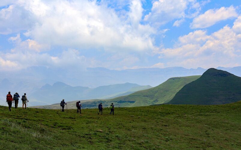 Un groupe de sept randonneurs portant des sacs à dos traverse une colline herbeuse au Lesotho, avec en arrière-plan des montagnes verdoyantes et un ciel partiellement nuageux. - Karavaniers