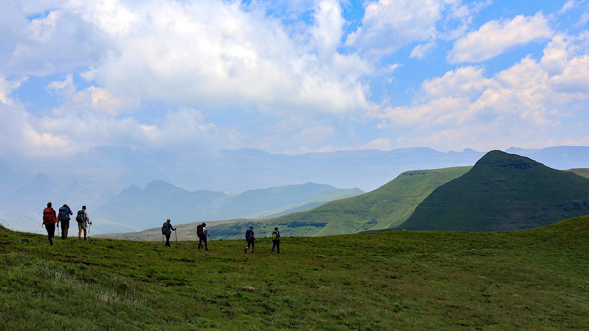Un groupe de sept randonneurs portant des sacs à dos traverse une colline herbeuse au Lesotho, avec en arrière-plan des montagnes verdoyantes et un ciel partiellement nuageux. - Karavaniers