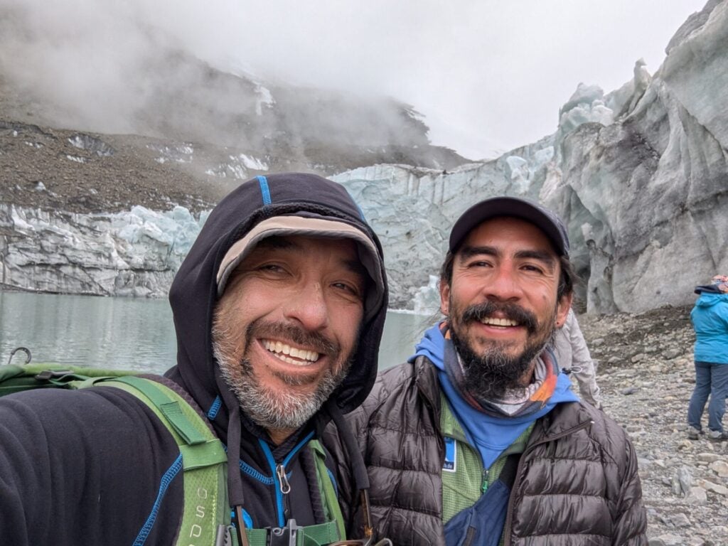 Deux hommes vêtus de vestes d'extérieur et de sacs à dos sourient pour un selfie devant un glacier ; des montagnes brumeuses, Javier Tellaeche à leurs côtés, un lac glaciaire et d'autres randonneurs complètent la toile de fond à couper le souffle. - Karavaniers