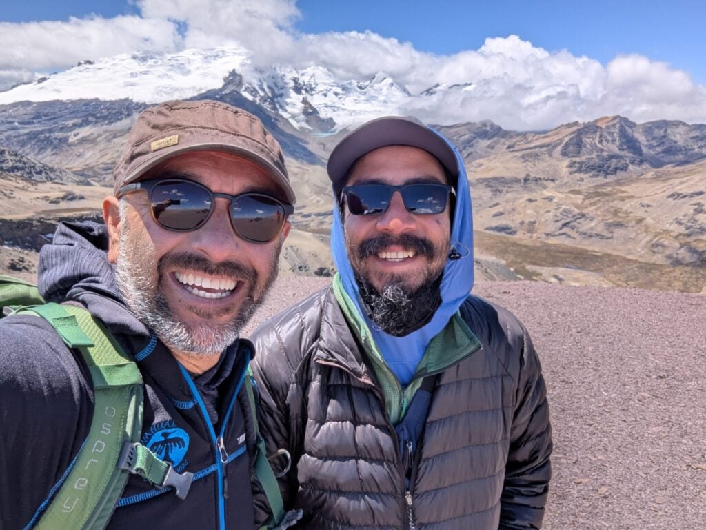 Deux hommes, dont Javier Tellaeche, portant des lunettes de soleil et des vestes d'extérieur sourient pour un selfie devant un paysage montagneux avec des sommets enneigés et un ciel partiellement nuageux en arrière-plan. - Karavaniers