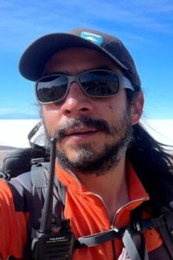 Cédric Blattner, cheveux longs, casquette, lunettes de soleil et veste orange, prend un selfie en plein air avec une radio accrochée à son sac à dos. En arrière-plan, on aperçoit un vaste paysage et un ciel d'un bleu éclatant. - Karavaniers