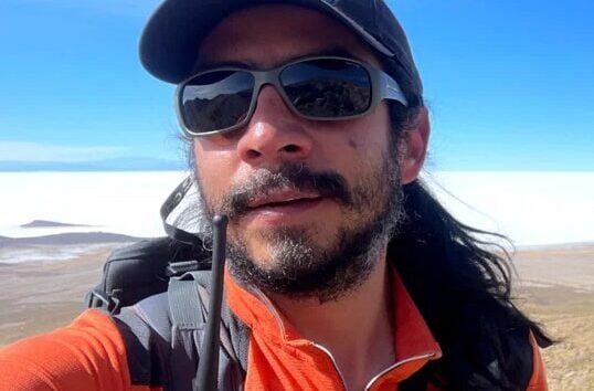 Cédric Blattner, cheveux longs, casquette, lunettes de soleil et veste orange, prend un selfie en plein air avec une radio accrochée à son sac à dos. En arrière-plan, on aperçoit un vaste paysage et un ciel d'un bleu éclatant. - Karavaniers