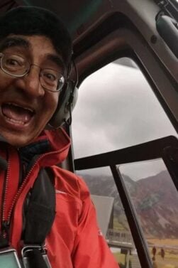 Cédric Blattner, vêtu d'une veste rouge et d'un casque, sourit et lève le pouce alors qu'il est assis à l'intérieur d'un hélicoptère. Par la porte ouverte, on aperçoit des montagnes et un ciel nuageux. - Karavaniers