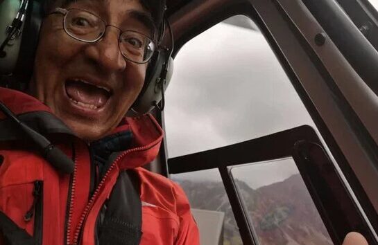 Cédric Blattner, vêtu d'une veste rouge et d'un casque, sourit et lève le pouce alors qu'il est assis à l'intérieur d'un hélicoptère. Par la porte ouverte, on aperçoit des montagnes et un ciel nuageux. - Karavaniers