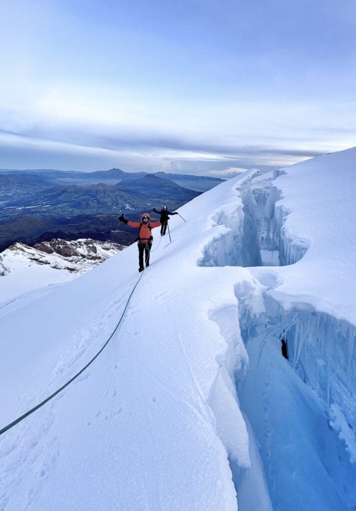 Deux alpinistes encordés marchent le long d'une arête enneigée avec de profondes crevasses sur un glacier, entouré de montagnes sous un ciel nuageux. - Karavaniers