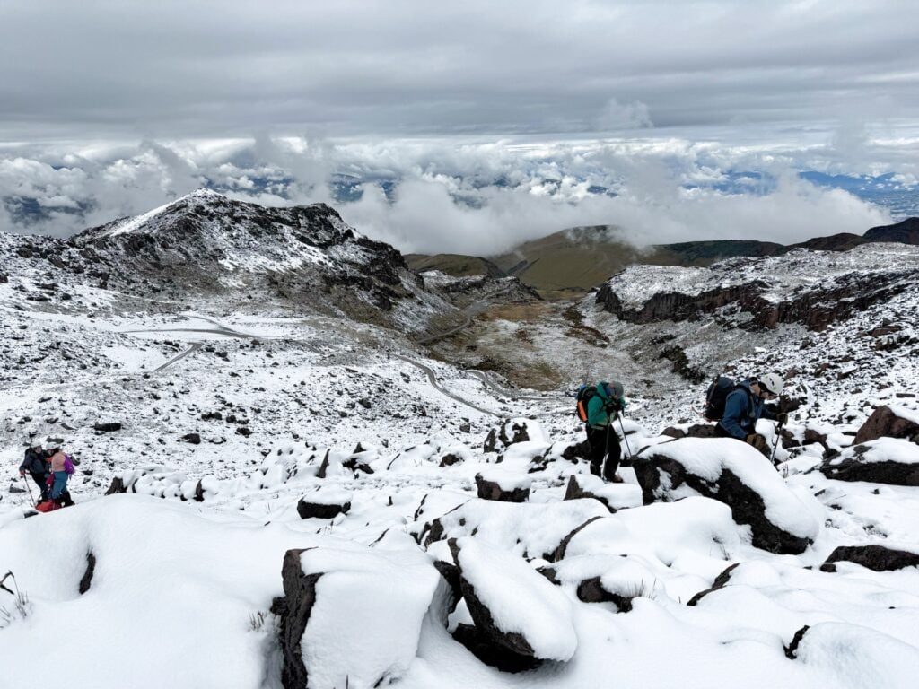 Plusieurs randonneurs portant des sacs à dos progressent sur un versant rocheux et enneigé d'une montagne sous un ciel nuageux. Au loin, on aperçoit des collines ondulantes et une couverture nuageuse. - Karavaniers