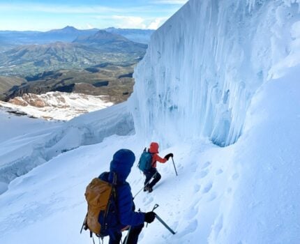 Deux alpinistes munis de sacs à dos et de piolets gravissent une pente raide et enneigée à côté d'un grand mur de glace sur une montagne, reliés par une corde. Des montagnes et des vallées sont visibles à l'arrière-plan sous un ciel partiellement nuageux. - Karavaniers