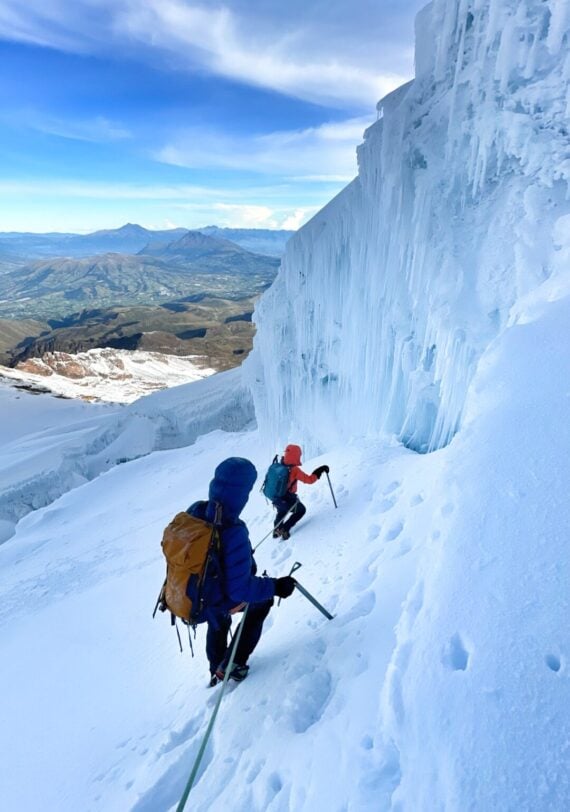 Deux alpinistes munis de sacs à dos et de piolets gravissent une pente raide et enneigée à côté d'un grand mur de glace sur une montagne, reliés par une corde. Des montagnes et des vallées sont visibles à l'arrière-plan sous un ciel partiellement nuageux. - Karavaniers