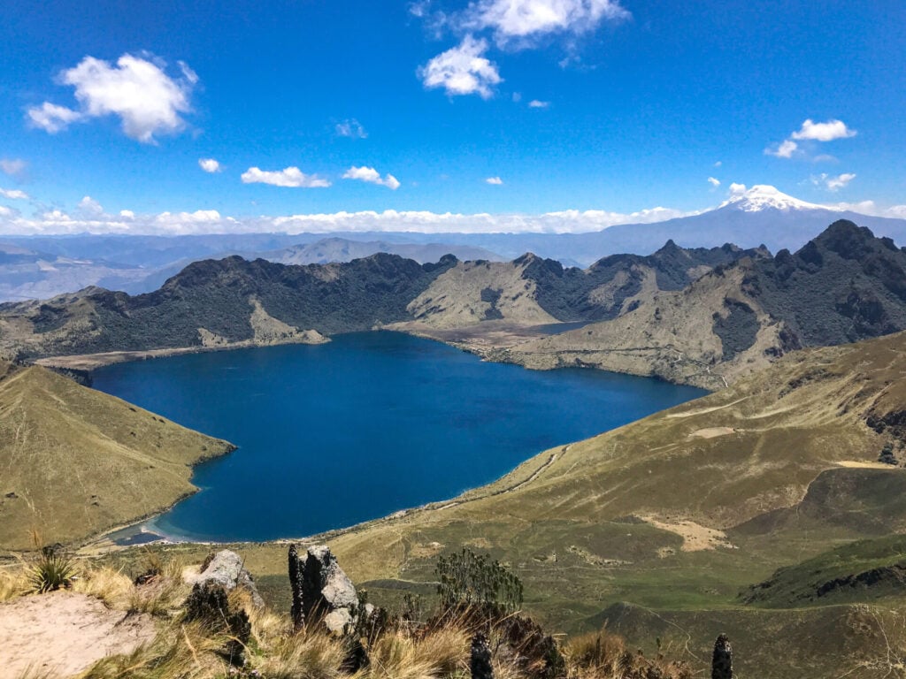 Un grand lac de montagne d'un bleu profond, entouré de collines herbeuses et de pics escarpés, sous un ciel d'un bleu éclatant avec des nuages épars - une scène parfaite pour une randonnée sur mesure Équateur, avec des montagnes enneigées visibles à l'arrière-plan lointain. - Karavaniers