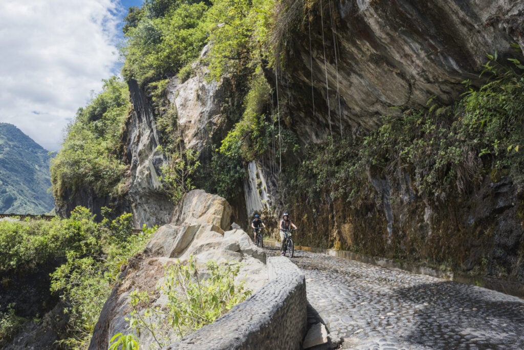 Deux cyclistes roulent sur une étroite route pavée creusée dans un flanc de montagne rocheux et escarpé, entouré d'une verdure luxuriante - une aventure qui rappelle un trek sur mesure Équateur sous un ciel partiellement nuageux. - Karavaniers