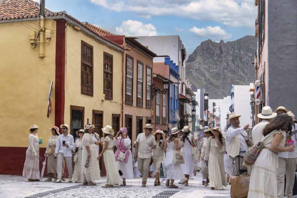 Un groupe de personnes vêtues de vêtements blancs de style ancien marche le long d'une rue pavée bordée de bâtiments colorés, avec des montagnes visibles en arrière-plan sous un ciel partiellement nuageux - rappelant une aventure de randonnée aux Îles Canaries. - Karavaniers