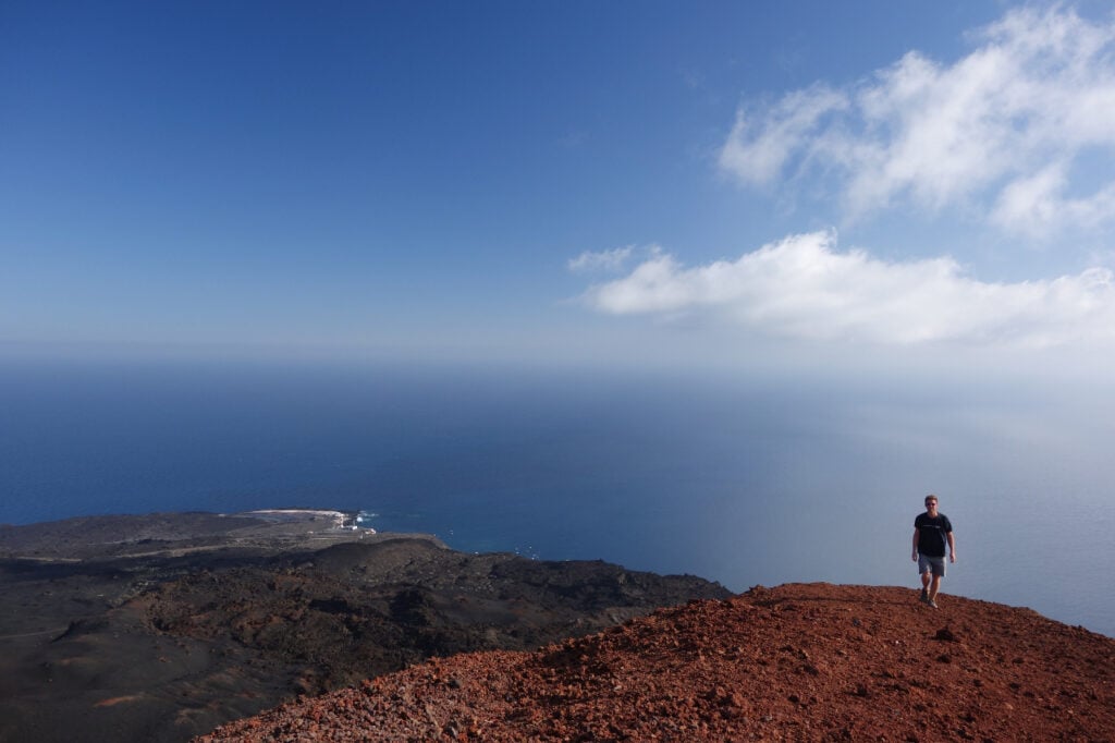 Une personne se trouve au sommet d'une colline rocheuse rougeâtre, surplombant un vaste océan bleu et une côte lointaine, sous un ciel clair avec quelques nuages. - Karavaniers