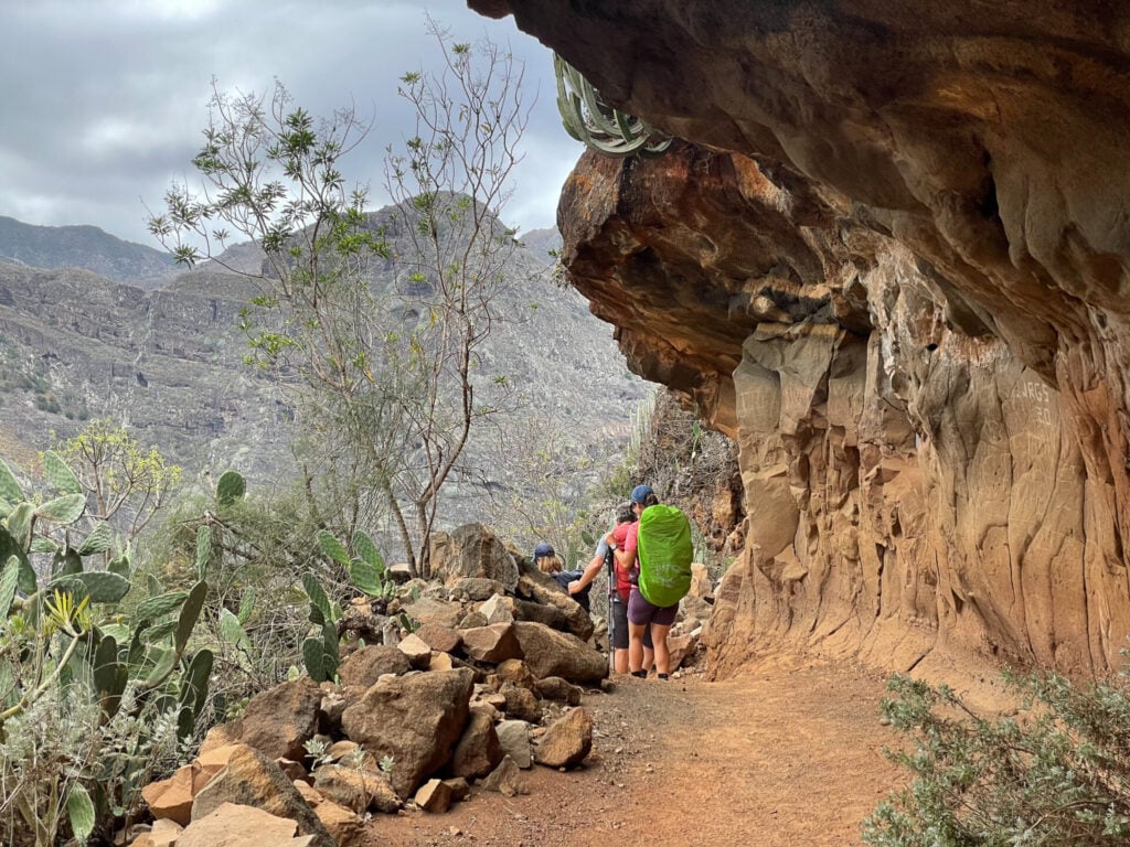 Trois randonneurs avec des sacs à dos se tiennent sur un sentier rocailleux sous une falaise en surplomb, entourés de cactus et d'arbustes, avec des montagnes en arrière-plan sous un ciel nuageux. - Karavaniers