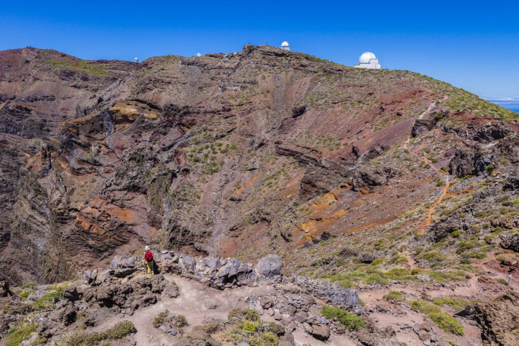 Un randonneur se tient sur un sentier rocailleux dans un paysage montagneux au sol brun-rouge et à la végétation clairsemée. Plusieurs dômes d'observatoires sont visibles sur la crête sous un ciel bleu clair. - Karavaniers