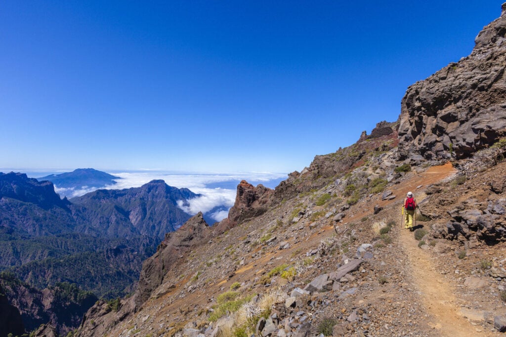 Un randonneur portant un sac à dos rouge marche sur un sentier de montagne rocailleux aux pentes abruptes. Au loin, des montagnes boisées s'élèvent au-dessus d'une couche de nuages dans un ciel bleu clair. - Karavaniers