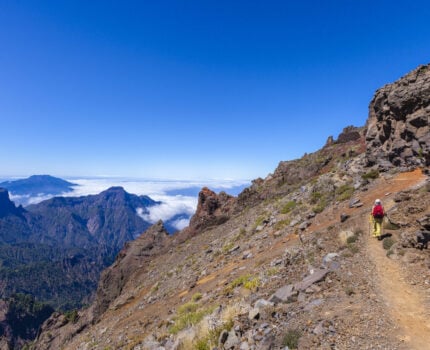 Un randonneur portant un sac à dos rouge marche sur un sentier de montagne rocailleux aux pentes abruptes. Au loin, des montagnes boisées s'élèvent au-dessus d'une couche de nuages dans un ciel bleu clair. - Karavaniers