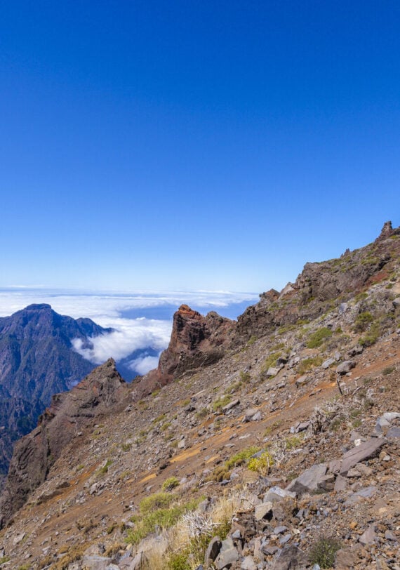 Un randonneur portant un sac à dos rouge marche sur un sentier de montagne rocailleux aux pentes abruptes. Au loin, des montagnes boisées s'élèvent au-dessus d'une couche de nuages dans un ciel bleu clair. - Karavaniers