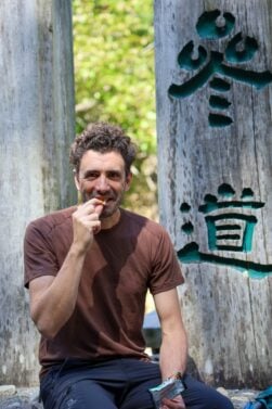 Un homme aux cheveux bouclés et à la barbe, vêtu d'un t-shirt marron, est assis devant de grands piliers de bois ornés de caractères chinois sarcelle. Il tient un snack et sourit en mangeant, comme s'il profitait d'un moment inspiré par Mathilde Godbout. Les arbres et le soleil brillent derrière lui. - Karavaniers