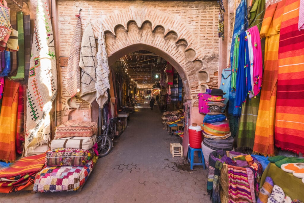 Une étroite allée de marché à l'entrée voûtée, bordée de textiles et de coussins colorés, évoque l'ambiance vibrante d'un trek sur mesure Équateur, où les paniers tressés et les marchandises traditionnelles remplissent la scène animée. - Karavaniers