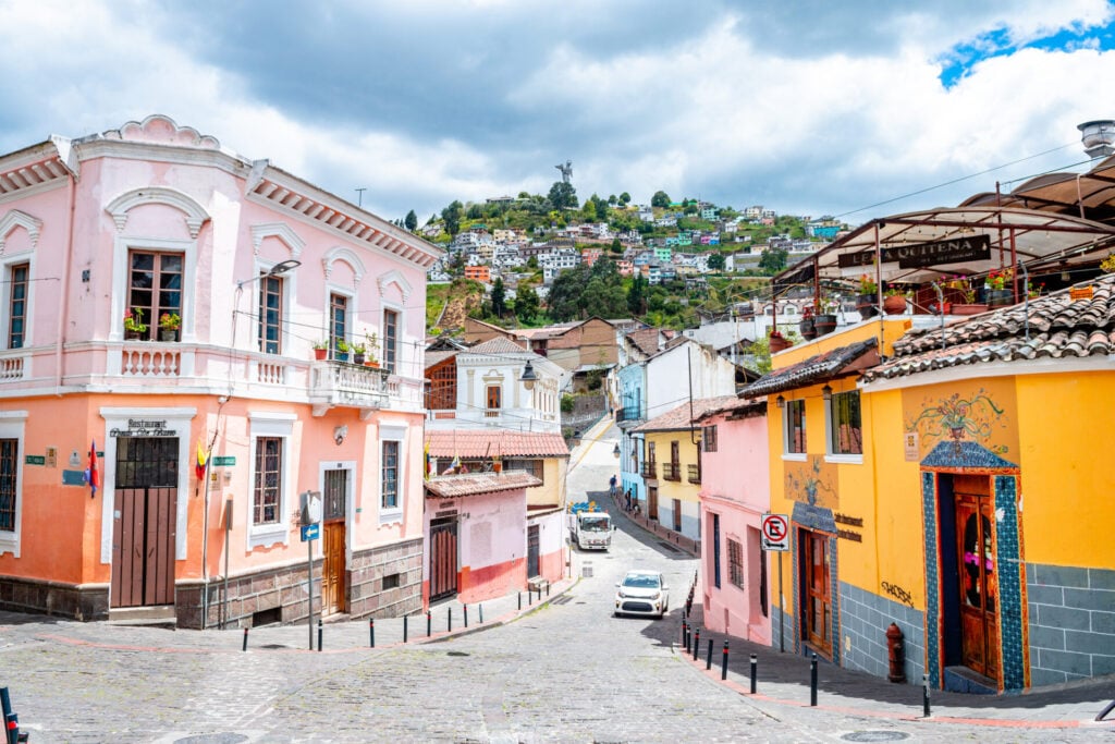 Une rue pavée dans une ville historique bordée de bâtiments colorés ; un bâtiment rose à gauche, un bâtiment jaune à droite, et des maisons sur une colline avec une grande statue visible à l'arrière-plan sous un ciel nuageux. - Karavaniers
