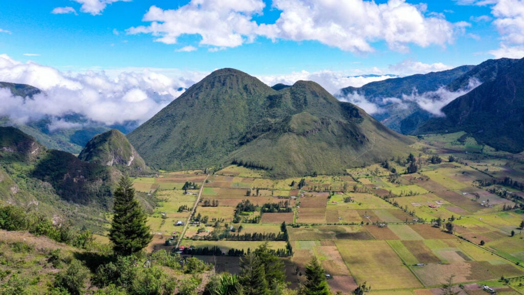 Vue aérienne d'une vallée verdoyante avec des terres agricoles en patchwork et des maisons éparses, bordée de montagnes boisées sous un ciel bleu avec des nuages. - Karavaniers