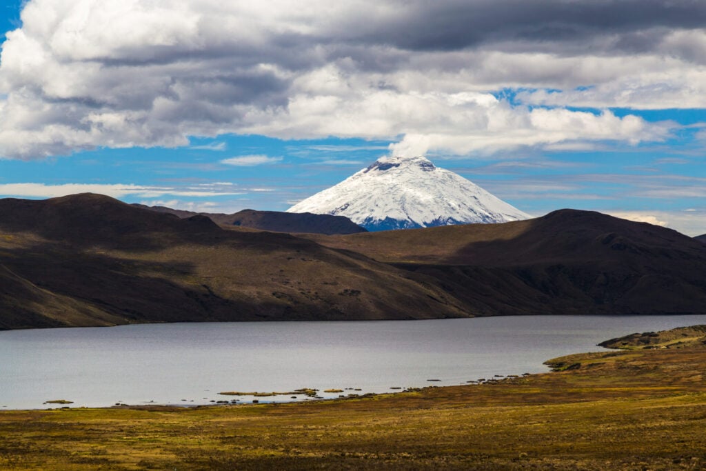 Un volcan enneigé se dresse à l'arrière-plan sous un ciel partiellement nuageux, avec des collines brunes et un lac calme au premier plan. - Karavaniers