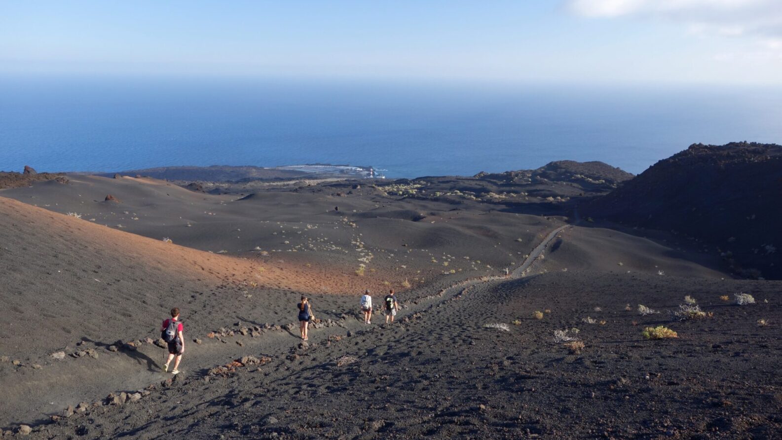 Un groupe de personnes descend une pente rocheuse et volcanique en direction de l'océan, tout en discutant des nouveautés en matière de géologie. Le paysage aride, au sol volcanique sombre et à la végétation clairsemée, s'étend sous un ciel clair parsemé de nuages près de l'horizon. - Karavaniers