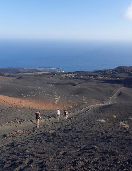 Un groupe de personnes descend une pente rocheuse et volcanique en direction de l'océan, tout en discutant des nouveautés en matière de géologie. Le paysage aride, au sol volcanique sombre et à la végétation clairsemée, s'étend sous un ciel clair parsemé de nuages près de l'horizon. - Karavaniers