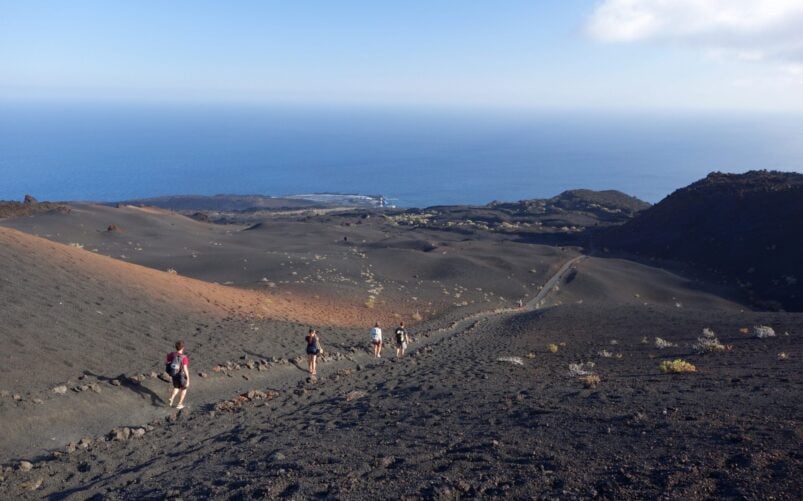 Un groupe de personnes descend une pente rocheuse et volcanique en direction de l'océan, tout en discutant des nouveautés en matière de géologie. Le paysage aride, au sol volcanique sombre et à la végétation clairsemée, s'étend sous un ciel clair parsemé de nuages près de l'horizon. - Karavaniers