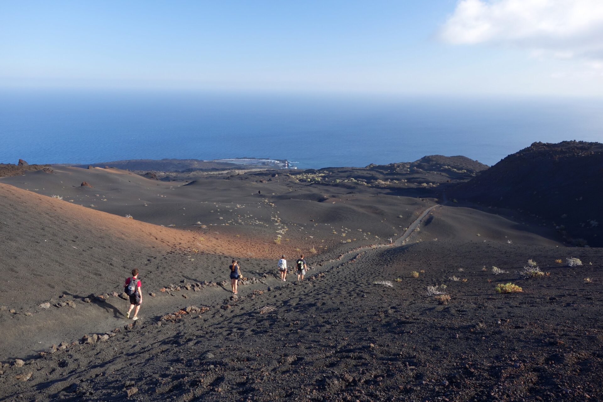 Un groupe de personnes descend une pente rocheuse et volcanique en direction de l'océan, tout en discutant des nouveautés en matière de géologie. Le paysage aride, au sol volcanique sombre et à la végétation clairsemée, s'étend sous un ciel clair parsemé de nuages près de l'horizon. - Karavaniers