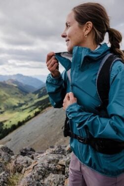 Caroline Côté, vêtue d'une veste bleue et d'un sac à dos noir, se tient sur un terrain rocailleux et ferme sa veste, avec en arrière-plan des collines et des montagnes verdoyantes s'étendant sous un ciel nuageux. - Karavaniers