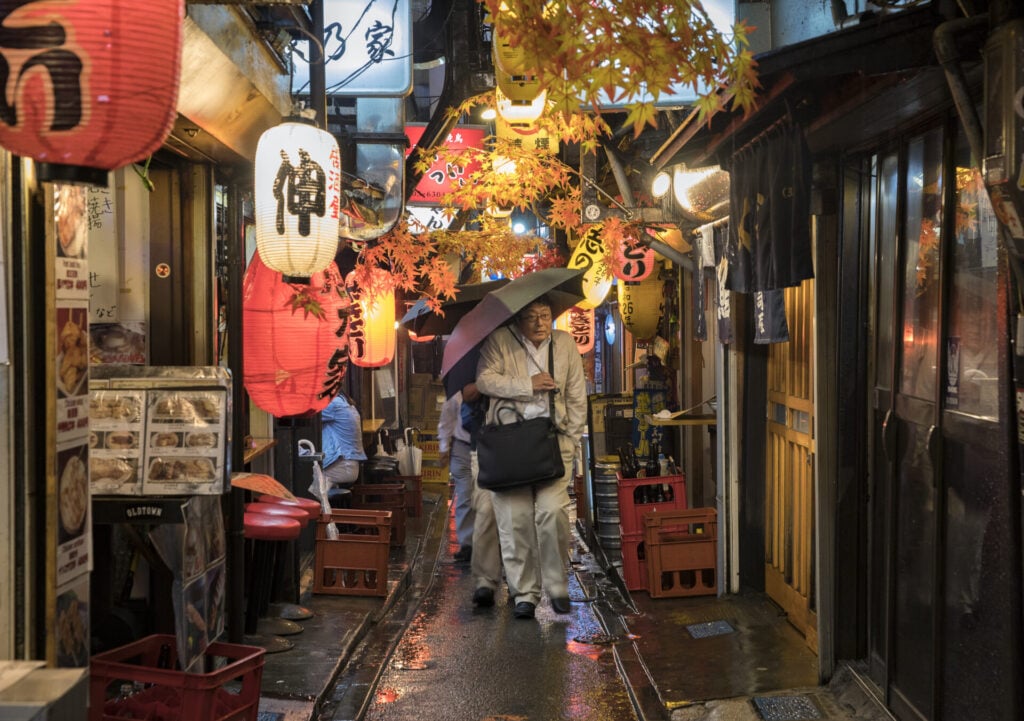 Une personne tenant un parapluie marche dans une ruelle étroite au sol mouillé, éclairée par des lanternes japonaises rouges et blanches qui rappellent une randonnée sur le Nakasendo, avec des enseignes de restaurants et des menus qui bordent les deux côtés de la ruelle. - Karavaniers