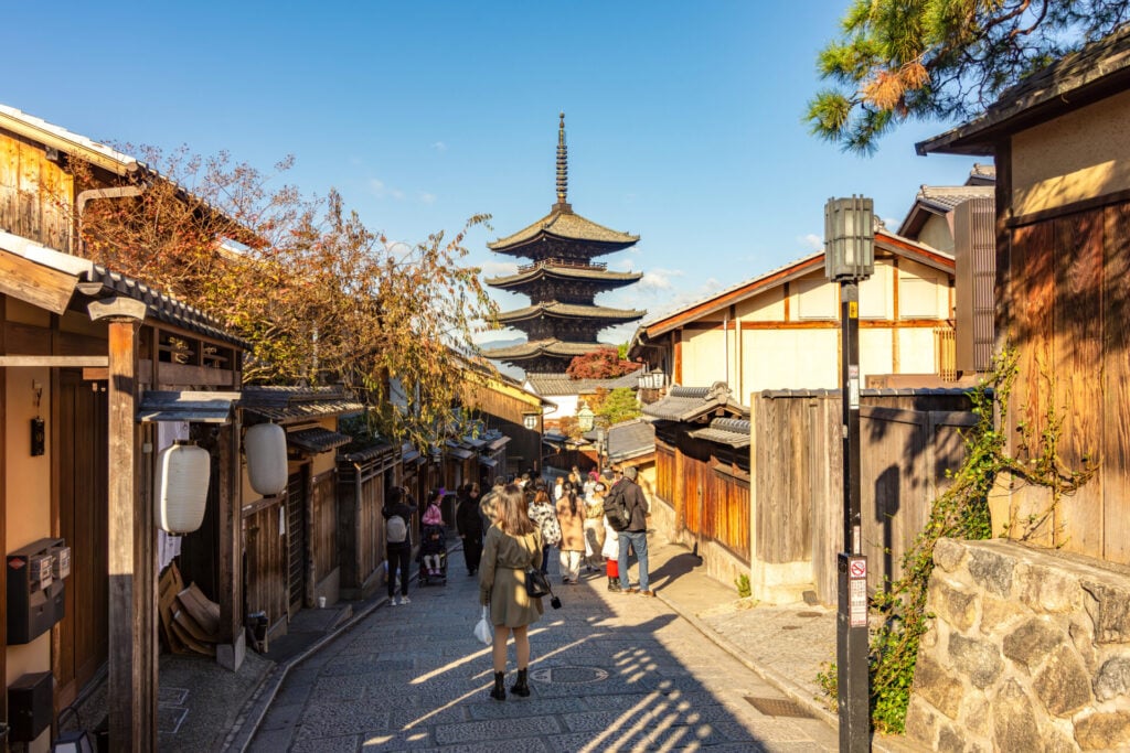 Scène de rue à Kyoto, au Japon, avec des personnes marchant le long d'un chemin de pierre bordé de bâtiments traditionnels en bois, et une grande pagode s'élevant à l'arrière-plan - faisant écho à la beauté calme que l'on trouve près des sommets sacrés des Alpes japonaises. - Karavaniers