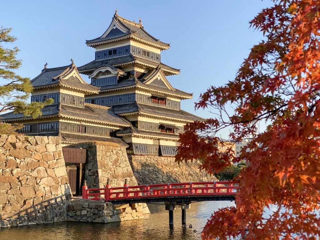Un château japonais traditionnel aux murs noirs et blancs se dresse à côté d'un mur de pierre et de douves, évoquant l'esprit d'une randonnée le long d'anciennes routes comme le Nakasendo. Un pont rouge et des feuilles d'automne brillent sous le ciel bleu clair. - Karavaniers