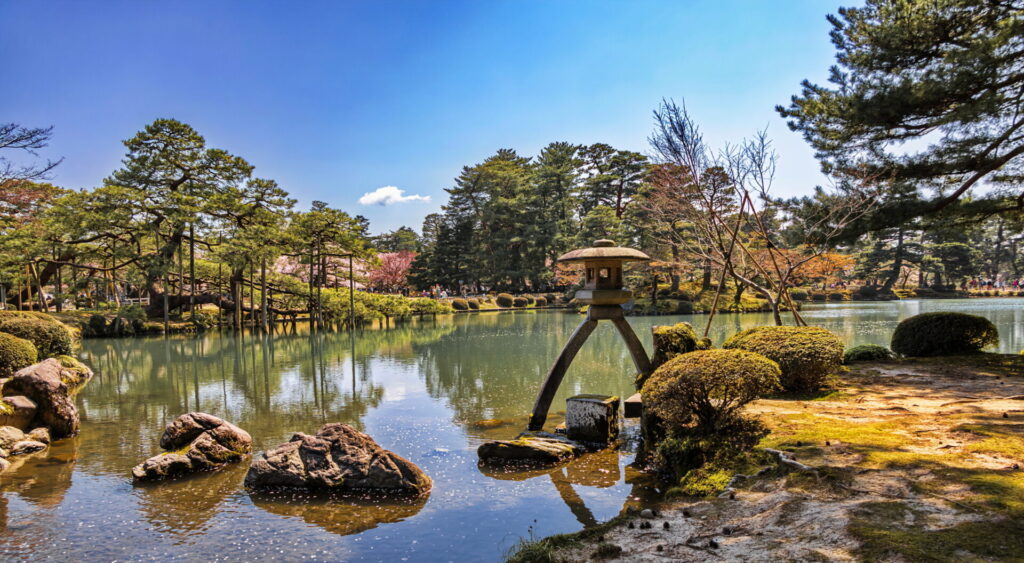 Un jardin japonais tranquille avec un étang, des rochers et une lanterne traditionnelle en pierre évoque la beauté sereine des Alpes japonaises. Le ciel bleu clair se reflète dans l'eau calme, tandis qu'une verdure luxuriante entoure cette retraite paisible. - Karavaniers