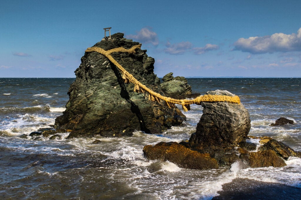 Deux gros rochers émergent de la mer, reliés par une corde épaisse. Les vagues s'écrasent autour d'eux. Une petite structure en bois, semblable à un portail, couronne le plus gros rocher. Une scène sereine qui rappelle une randonnée sur les anciennes routes japonaises du Kumano Kodo ou du Nakasendo. - Karavaniers