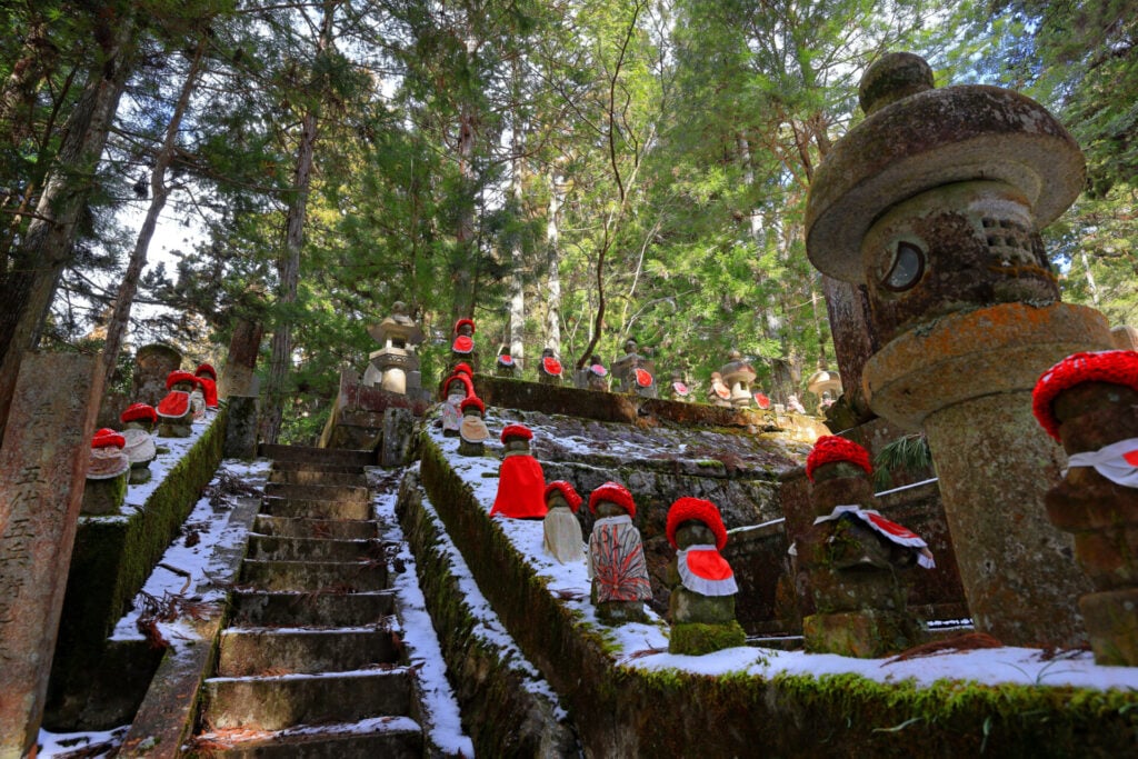 Des marches en pierre montent à travers une forêt moussue le long du sentier Kumano Kodo, flanquées de rangées de petites statues en pierre portant des bavoirs et des chapeaux rouges, avec des taches de neige sur le sol et des lanternes éparpillées. - Karavaniers