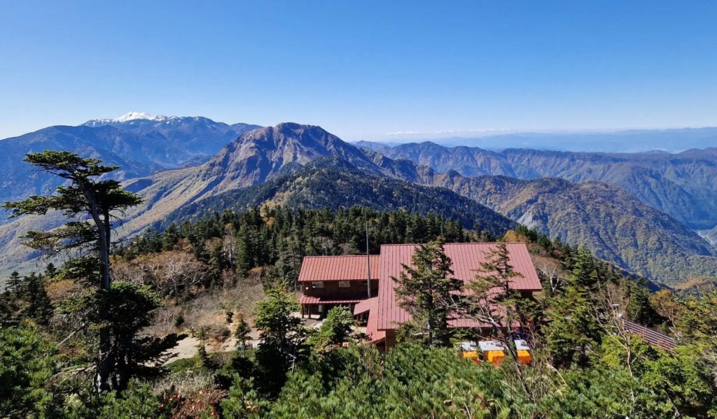 Un chalet de montagne aux toits rouges se dresse parmi les arbres verts sur une pente le long du légendaire trek de Kumano Kodo ; des montagnes boisées et escarpées s'étendent au loin sous un ciel bleu limpide. - Karavaniers